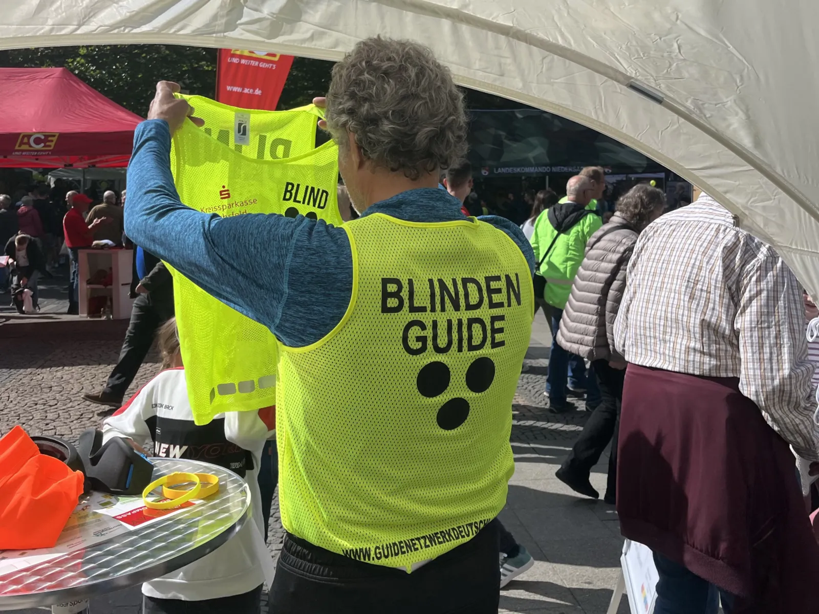 Ein Mann, der eine Weste auf der Aufschrift Blindenguide tr&auml;gt, eine weitere Weste h&auml;lt er in den H&auml;nden