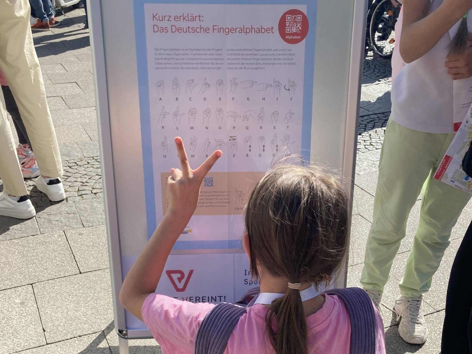 Ein Mädchen vor einem Plakat mit den Zeichen aus der Gebärdensprache, sie macht die Gebärde für V