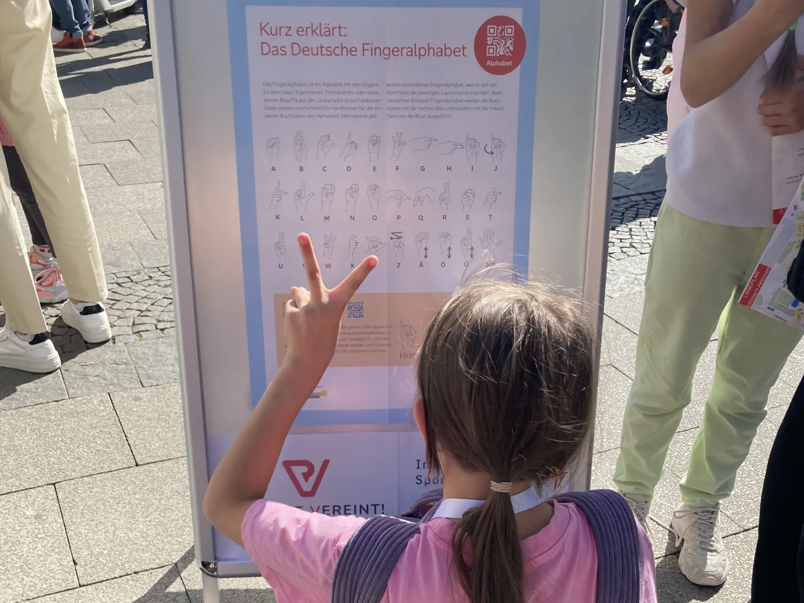 Ein M&auml;dchen vor einem Plakat mit den Zeichen aus der Geb&auml;rdensprache, sie macht die Geb&auml;rde f&uuml;r V